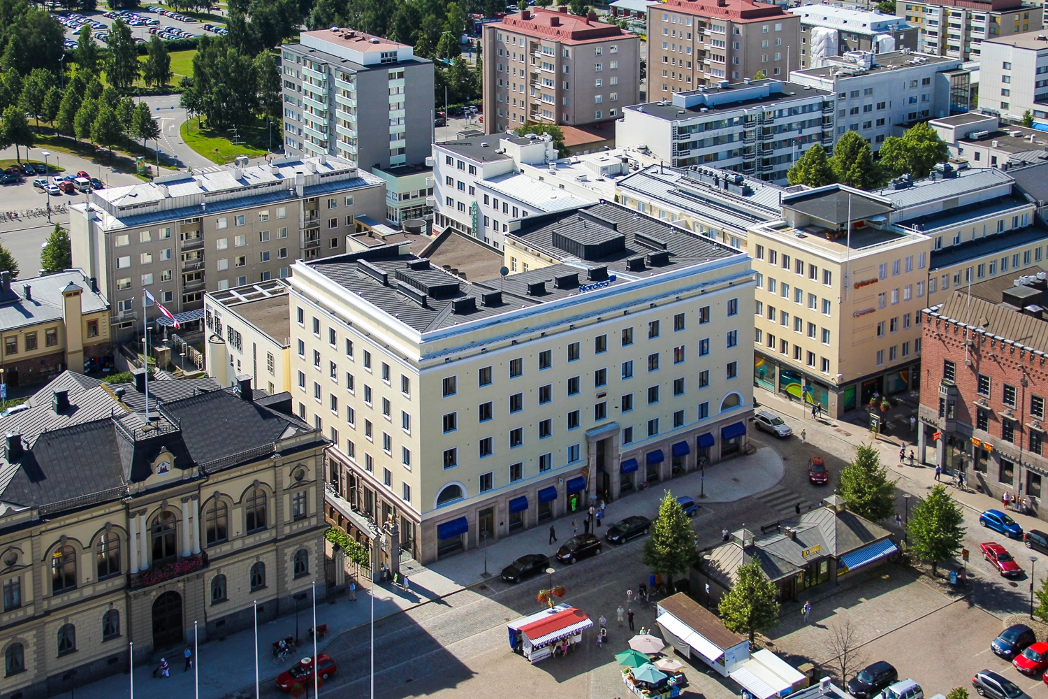 Hämeenlinnan Sibeliuksenkatu 2 ilmakuva. Kiinteistön julkisivu on vaaleankeltainen. Kiinteistön edessä on pieni toriaukeama jonka ympärillä on autoja pysäköitynä. Taustalla näkyy muita rakennuksia. 