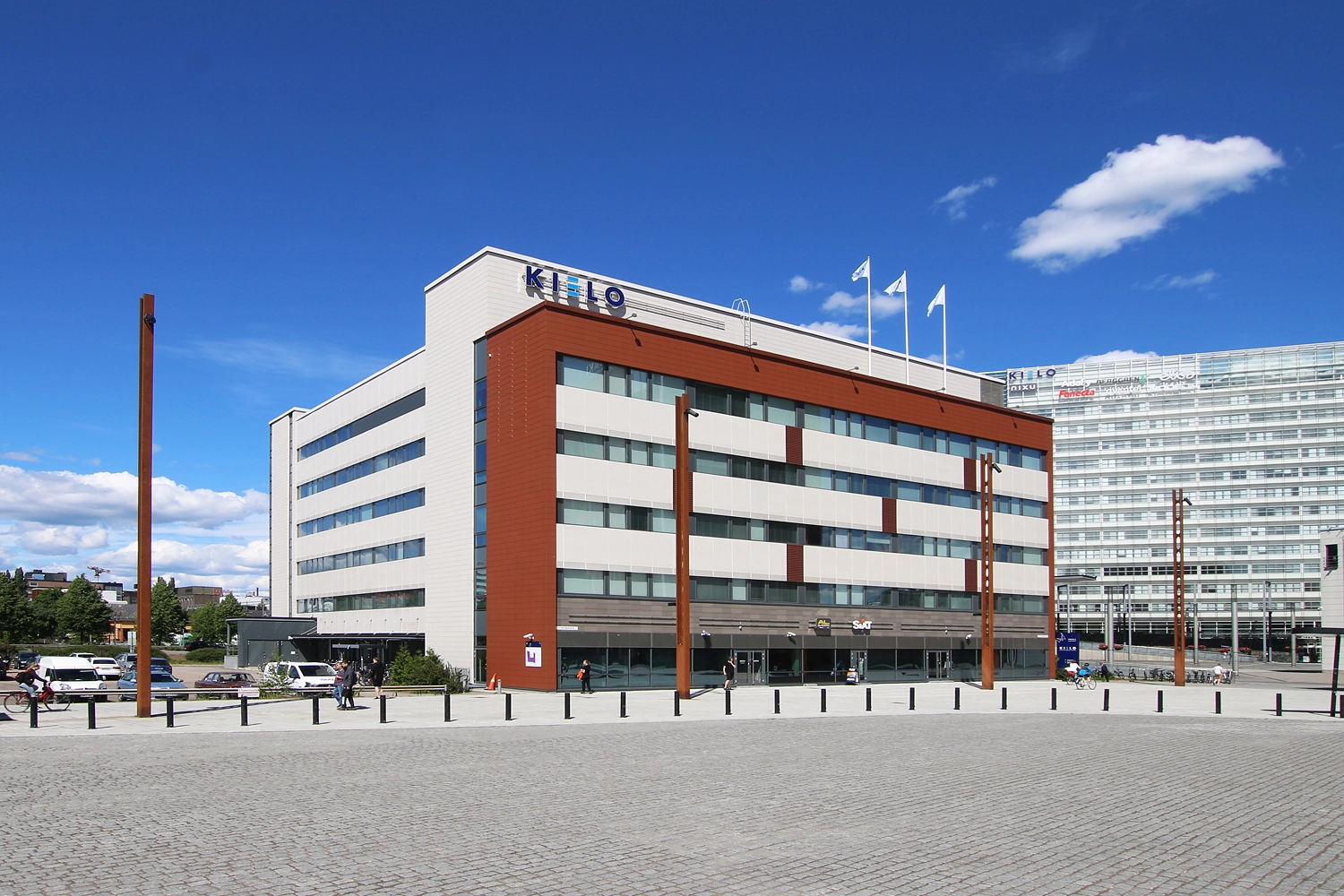 Innova 2, Lutakonaukio 7, Jyväskylä. Toimitilakiinteistön julkisivu on vaalea ja ruskea. Rakennuksen edessä kävelee ihmisiä kävelykadulla. Takana näkyy toinen Kielon rakennus