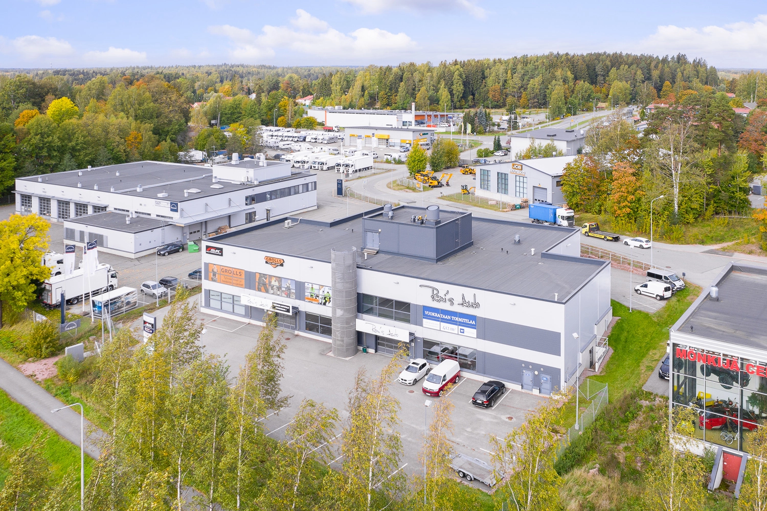 Espoon Kartanonherrantie 7, ilmakuva. Kaksikerroksinen, valkoinen ja harmaa rakennus jonka edustalla autoja pysäköitynä pysäköintialueelle. Ympärillä muita toimitilarakennuksia. Taustalla näkyy metsää